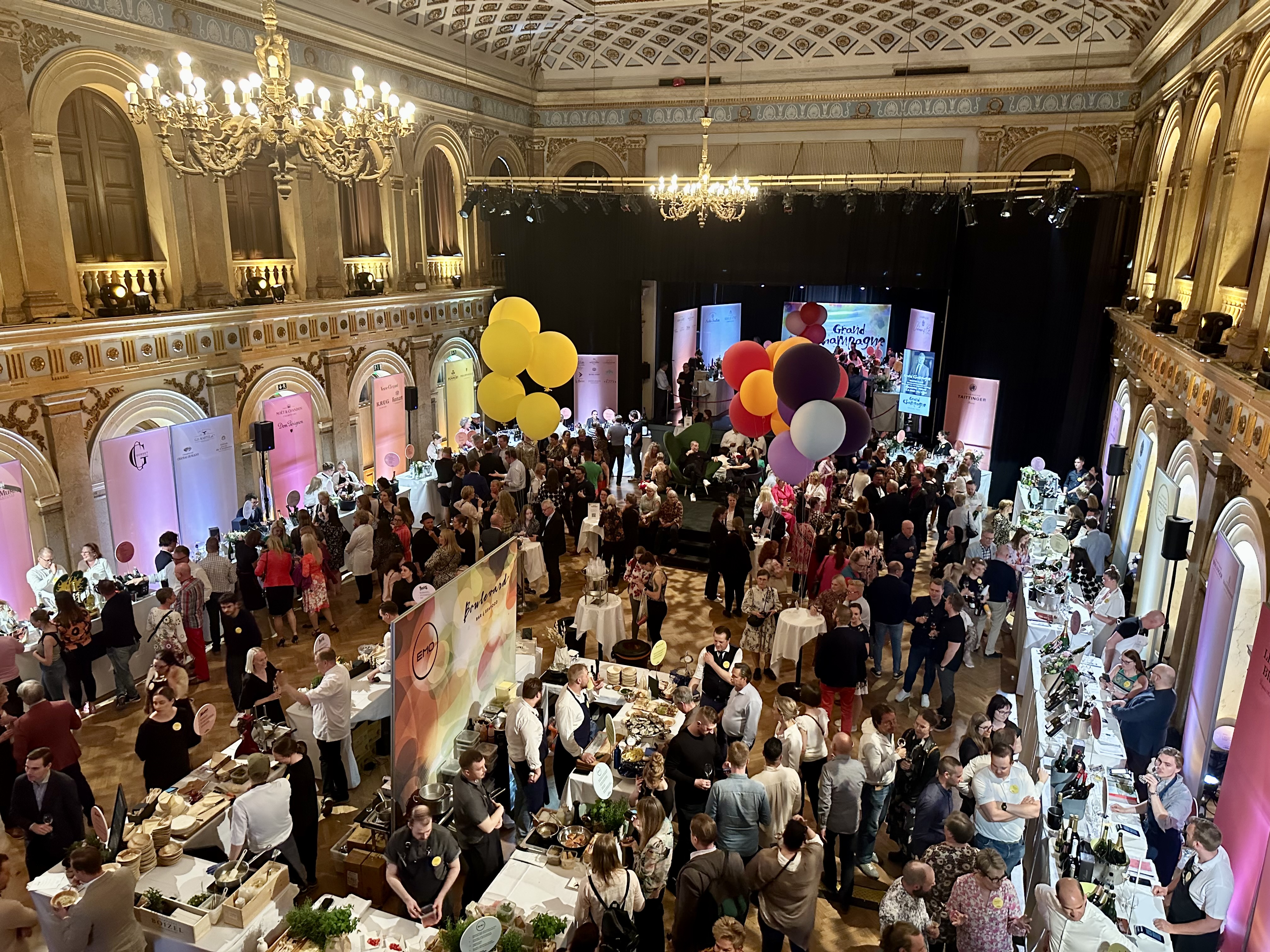 A bustling champagne tasting event with guests mingling in an ornate hall, featuring booths with champagne displays and large colorful balloons.