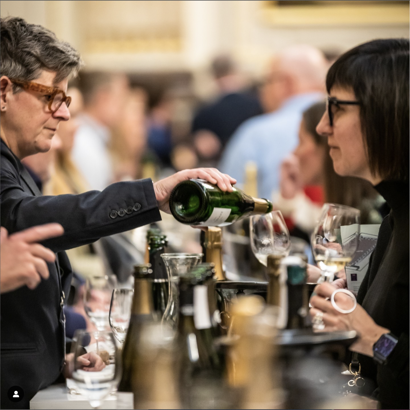 A woman pouring champagne at a tasting event, with guests engaged in conversation and various bottles of champagne displayed in the background.