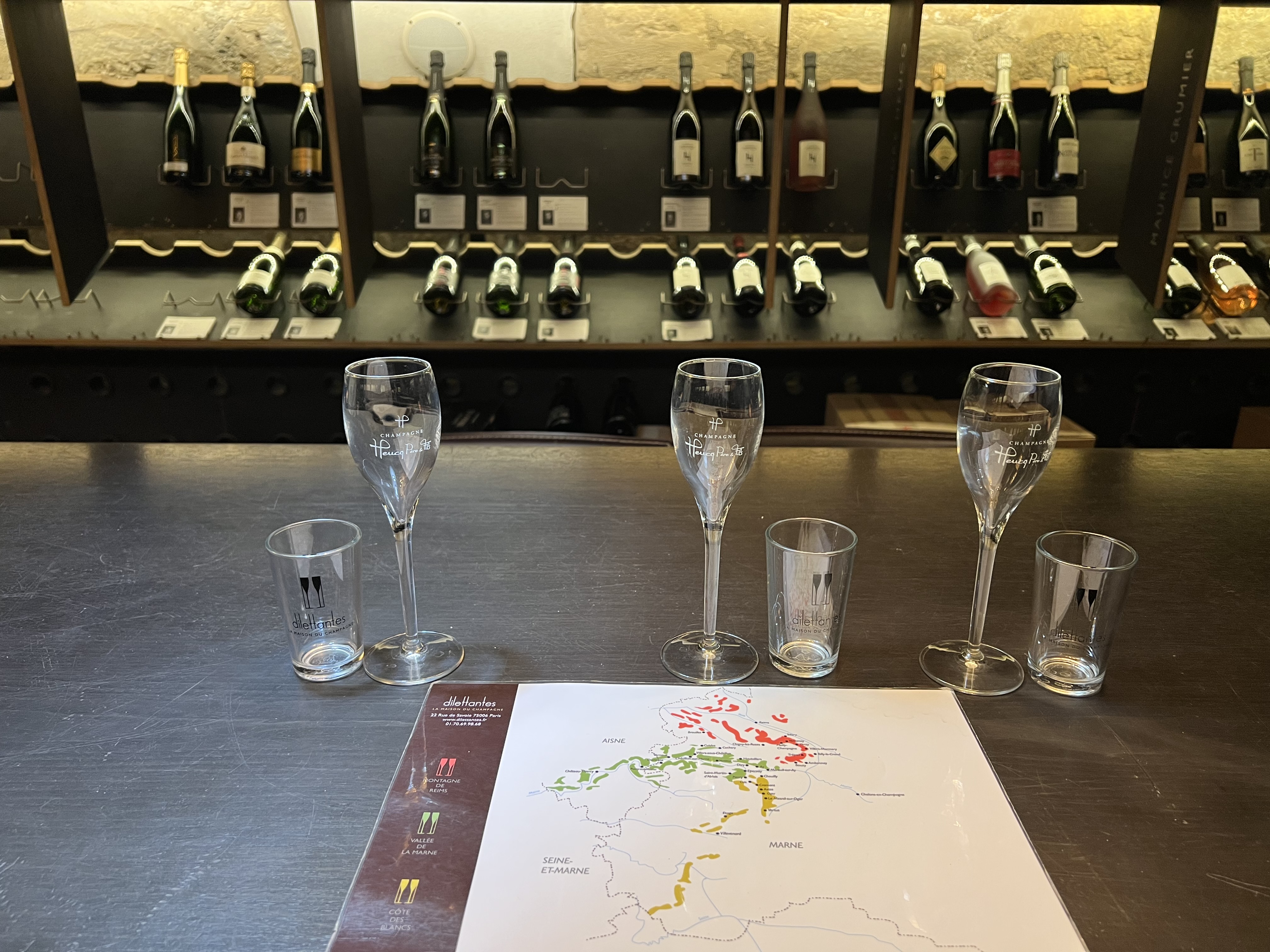 A bar counter displaying several champagne glasses and tasting vessels, with a region map and shelves of champagne bottles in the background.